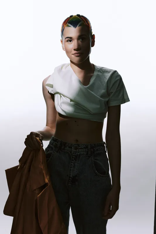 Young woman with colorful braided hair wearing casual white top and dark jeans holding a brown jacket, standing confidently against a bright neutral background.