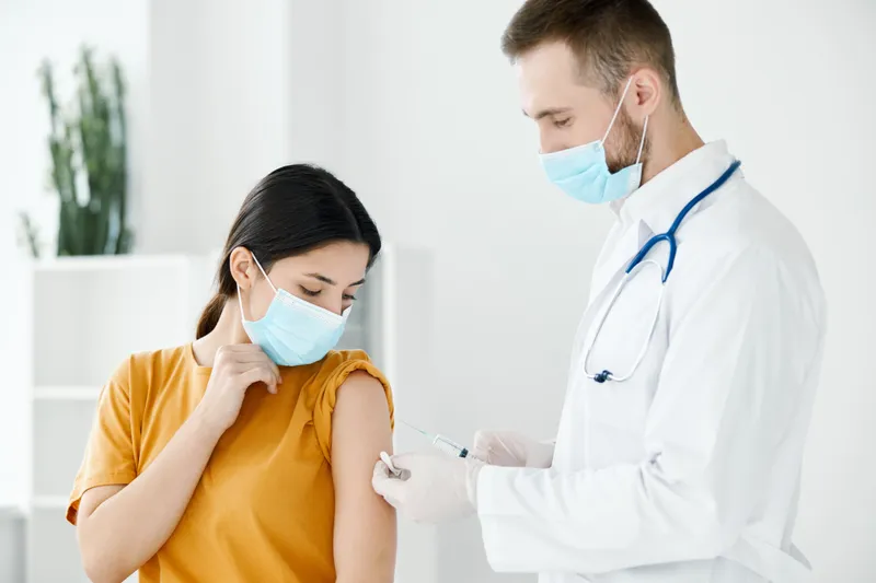 Врач делает женщине укол в плечо, на ней медицинская маска, вакцина от COVID-19