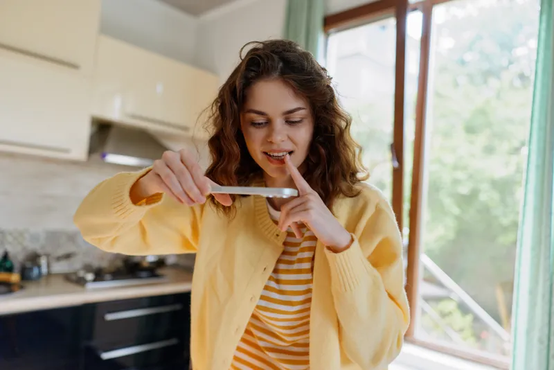 Cheerful woman indoors, examining a thermometer with curiosity, bright kitchen background, soft natural light, casual outfit, warm colors, relaxed pose, home life moment, everyday scene