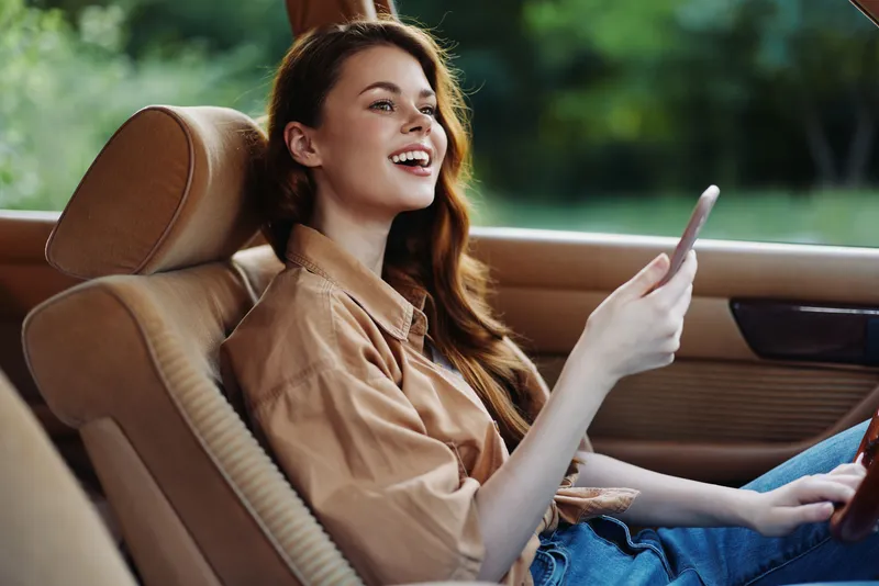 Young woman smiling while sitting in a car, enjoying a sunny day outdoors and using a smartphone, conveying joy and relaxation.