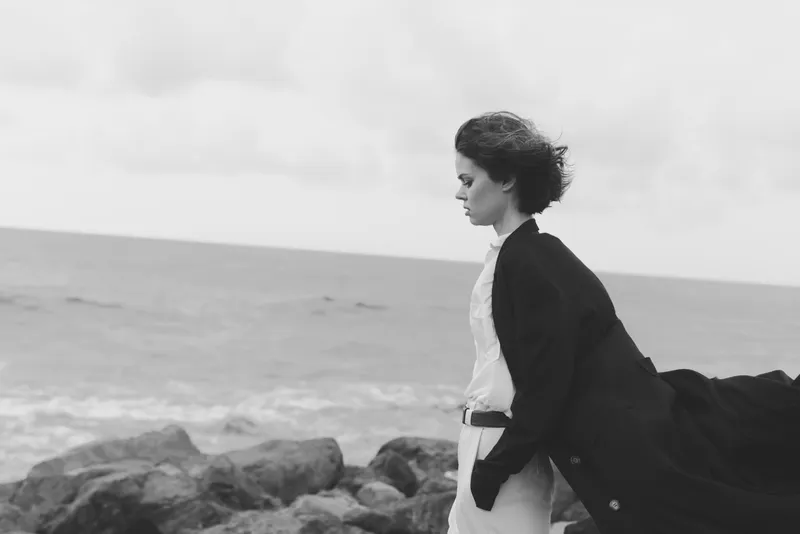Woman walks along a rocky coast as wind billows a coat, black and white scene capturing movement, sea spray, and quiet longing by the shore, dramatic shoreline silhouette