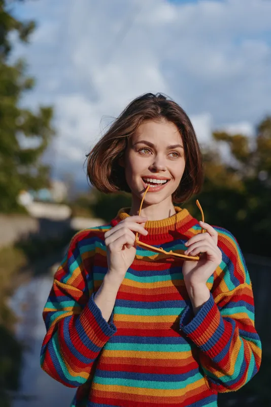 Woman smile sunglasses striped sweater portrait outdoors casual young adult holding sunglasses in colorful knitwear, short hair in natural daylight, happy expression and relaxed lifestyle vibe.