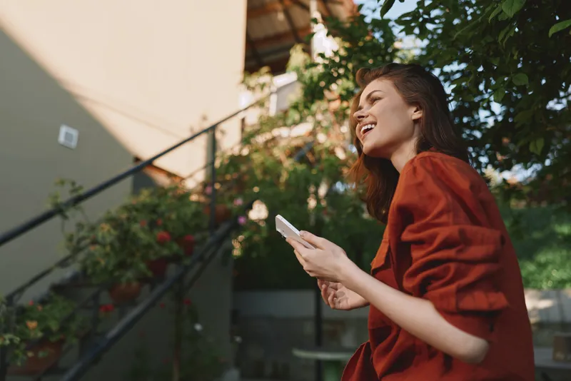 Woman outdoor lifestyle moment, smiling, casual red top, sunlit scene, phone in hand, relaxed posture, natural colors, greenery backdrop in a warm garden setting