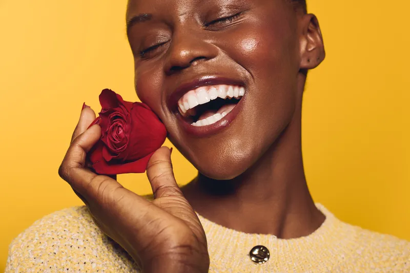 Happy African woman with dark skin smiles joyfully holding a red rose heart near her face on yellow background. Concept of love, happiness, skincare, and natural beauty with floral element.