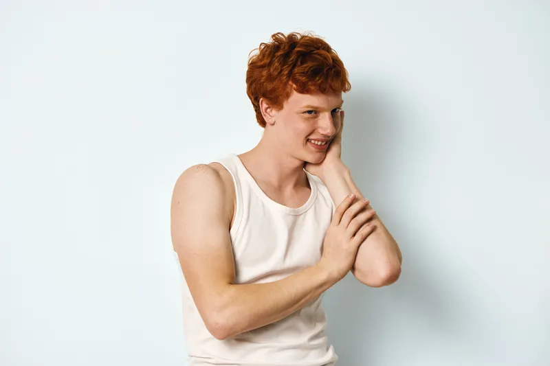 Young man with red hair wearing sleeveless white shirt posing against white background showing smile and casual relaxed posture. Male portrait with natural expression and light skin tone.