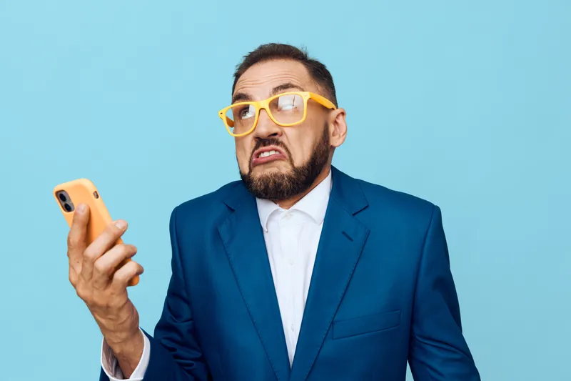 A business professional man in a blue suit on a solid colored background, expressing surprise and frustration while looking at a smartphone, conveying energy, tension, and focus in a studio shot.