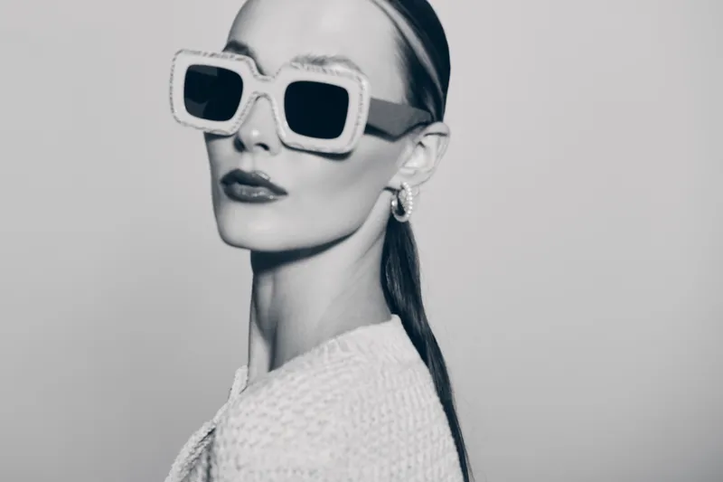 Stylish white woman with pale skin and slicked back hair wears large square sunglasses and textured clothing in monochrome studio portrait.