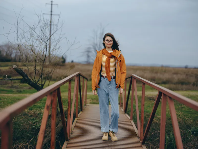 Woman walking on a wooden bridge outdoors in countryside lifestyle. Orange jacket, striped scarf, blue jeans. Overcast sky and cold air, natural path leading forward in autumn fields.