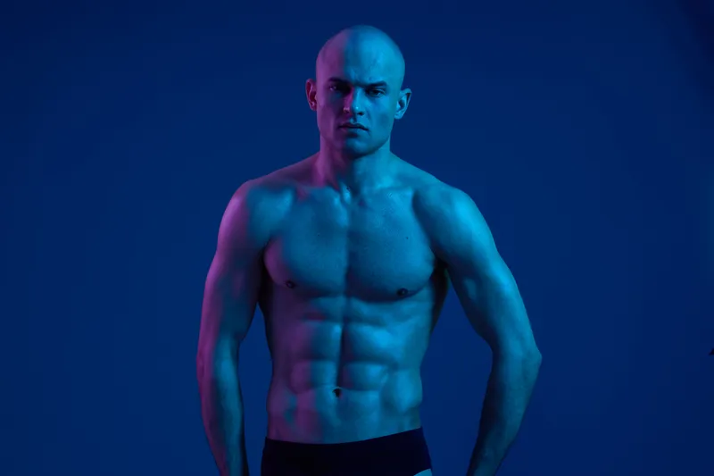 Athletic man posing confidently under blue lighting, showcasing his strong physique and focused expression, emphasizing power and determination.
