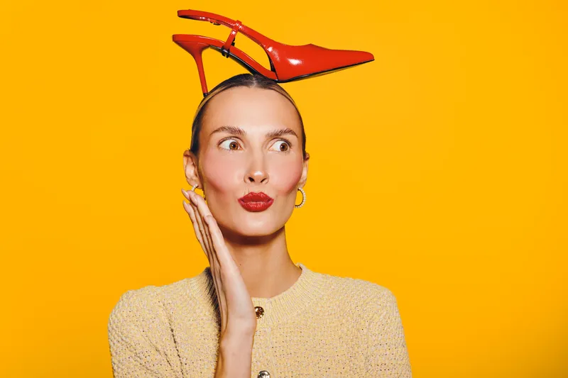 Young woman with red high heel shoe on head looks surprised on bright orange background. Concept of fashion, creativity, and playful style with bold colors and unique accessories.