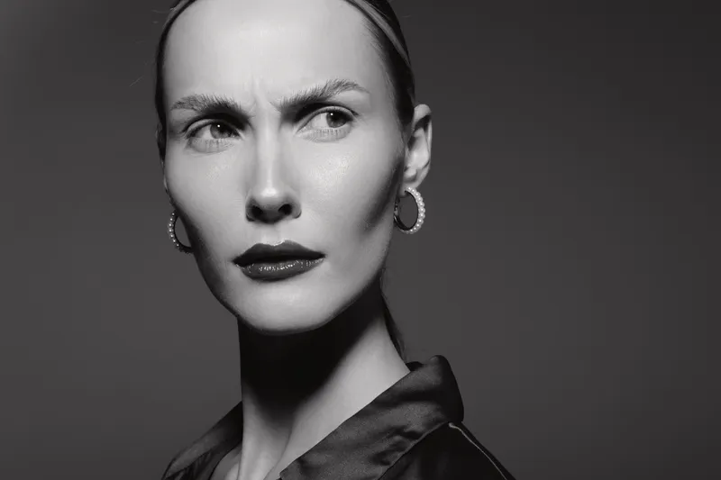 Portrait of a fair skinned white woman with slicked back hair and hoop earrings looking sideways with a serious expression in black and white studio lighting.