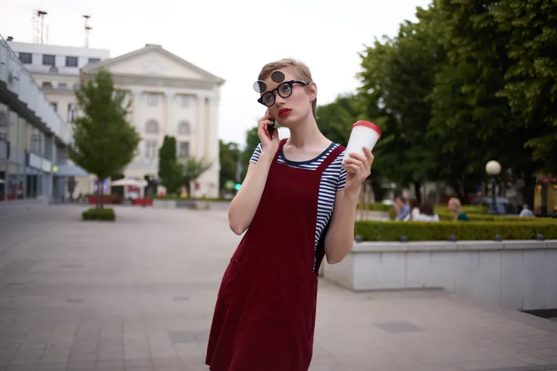 Женщина с короткой стрижкой на улице, в очках, со стаканом напитка в руке, идёт пешком