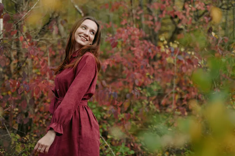 A candid portrait of a smiling young woman in a rust dress among autumn foliage, conveying warm authenticity and emotional reliability through natural pose, expression, and soft light.