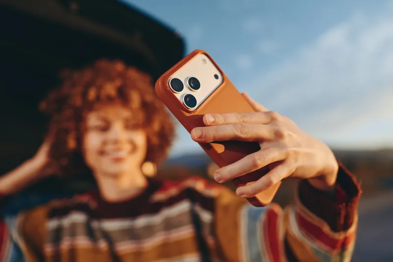 Woman holds smartphone for a selfie portrait with curly hair smiling in soft focus outdoors, closeup hand and orange phone case, lifestyle moment during summer daylight street walk