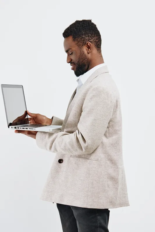 Confident smiling man with dark skin, wearing a light beige suit jacket and white shirt, holding a laptop and looking down, isolated on white background. Business casual, professional, modern style.