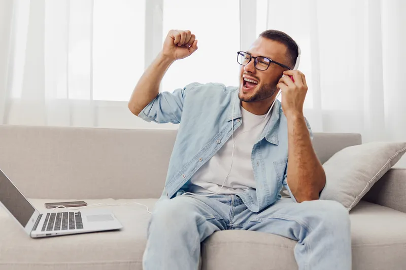 Energetic man enjoying music on headphones, sitting on a modern sofa with a laptop, celebrating his creative break. Perfect for showcasing relaxation and productivity.