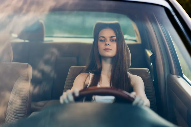 Young woman with long hair driving a car, focused and calm expression, in a sunlit interior, emphasizing freedom and independence, with a soft color palette.