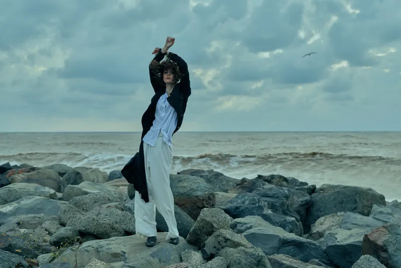 Fashionable figure stands on rocky shore by the sea, wind lifting a white dress and dark coat, dramatic pose amid clouds, ocean spray, and rugged coastline.