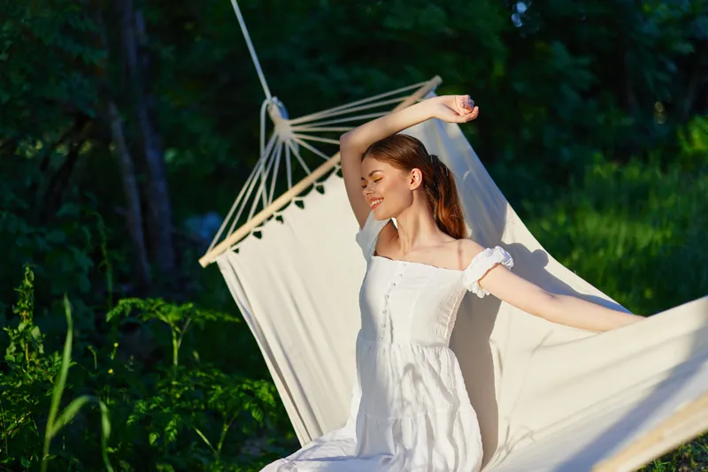 Young woman enjoying a moment of relaxation in a sunny garden, wearing a white dress, with a peaceful expression, surrounded by greenery