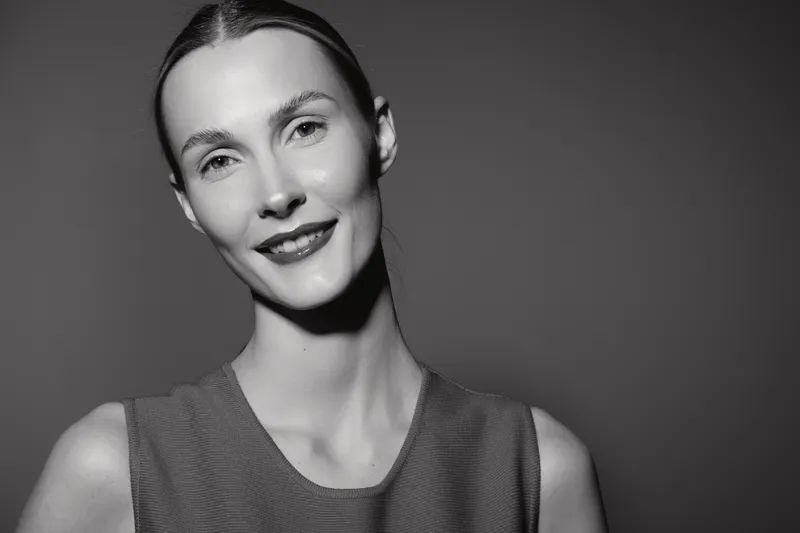 Close-up portrait of a smiling caucasian woman with fair skin and slicked-back hair against a dark background in black and white photography.