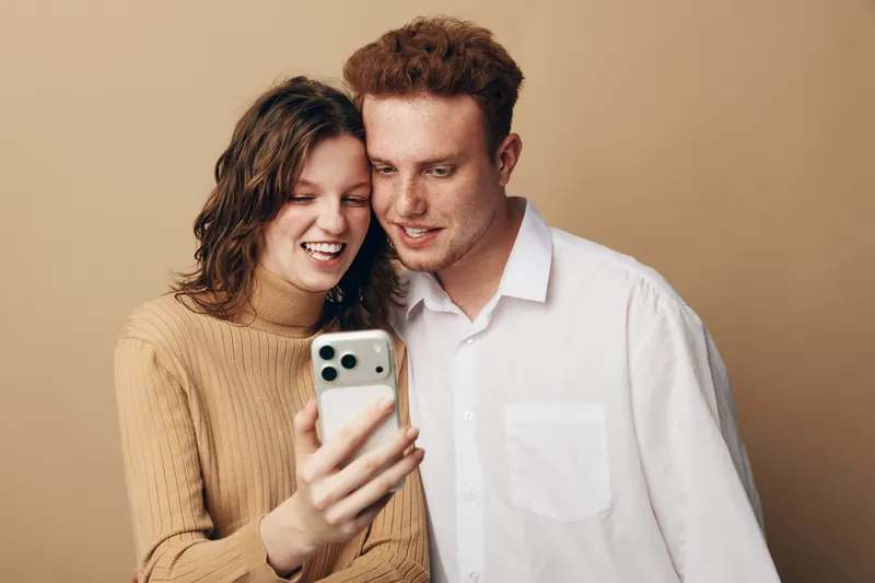 Young couple with casual clothes smiling and looking at smartphone screen together. Indoor studio portrait with warm beige background. Concept of technology, communication, and happiness.