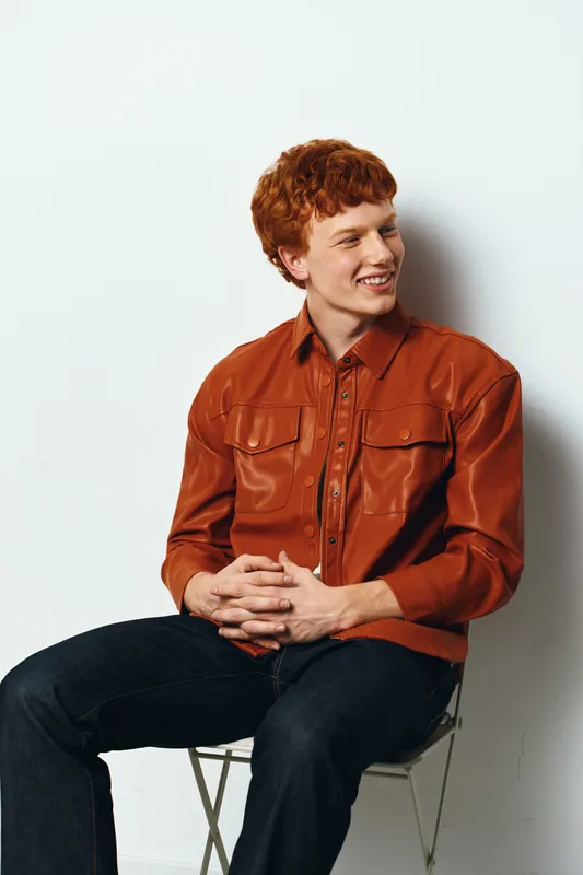 Young man with red hair wearing orange leather jacket and black pants sitting on folding chair against white background smiling and looking aside.