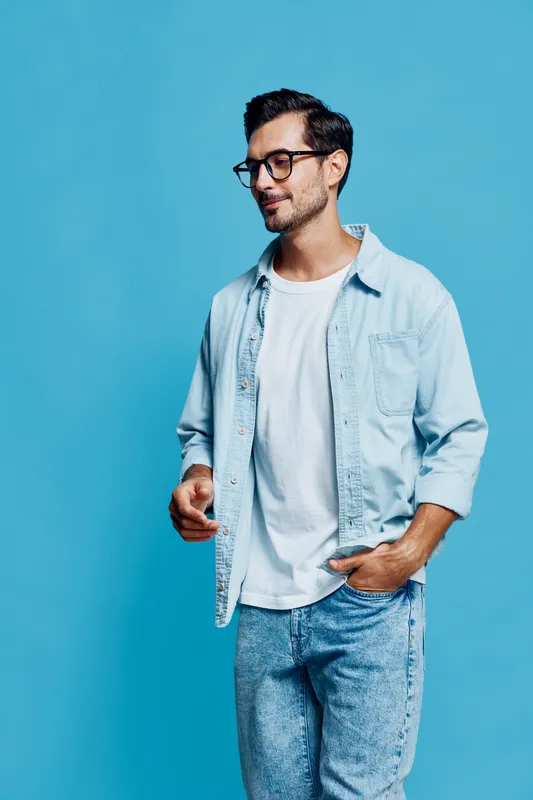 Stylish happy cheerful young man wearing glasses and denim shirt standing against a solid blue background, looking to the side with a confident expression.