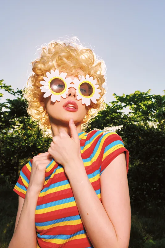 Whimsical young woman wears a curly blonde wig and daisy sunglasses, posing outdoors in a bright striped shirt with retro film colors and a playful mood.