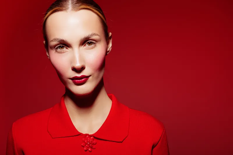 Portrait of a confident woman with red lipstick and red sweater on red background. Studio lighting highlights her facial features and smooth skin. Concept of fashion and beauty.