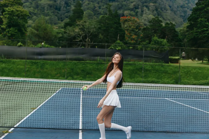 Young athletic woman playing tennis on a sunny day, wearing a white sporty outfit and demonstrating joy, set against a lush green background