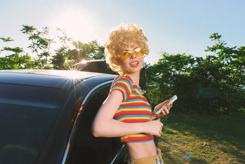 A woman with a retro hairstyle in a striped top stands by a car in bright sunlight, showcasing casual outdoor fashion and a vintage lifestyle moment.