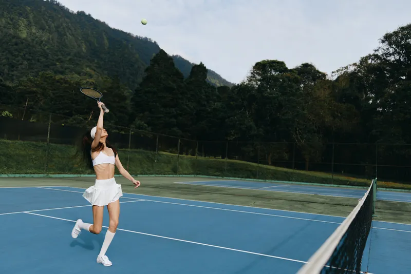 Young athletic woman playing tennis in a white sports outfit on a blue court, showcasing determination against a natural mountain backdrop