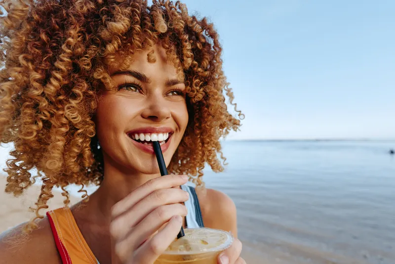 Curly haired young woman smiles while sipping a cold drink on a sunny beach, enjoying a carefree summer moment, with clear blue water in the background and warm light enhancing her glow.
