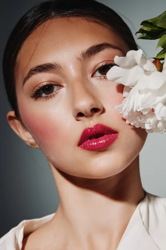 Close-up of a young woman with glossy red lipstick and bold makeup holding a white flower. Studio shot emphasizes fashion beauty, clean skin, and creative expression.