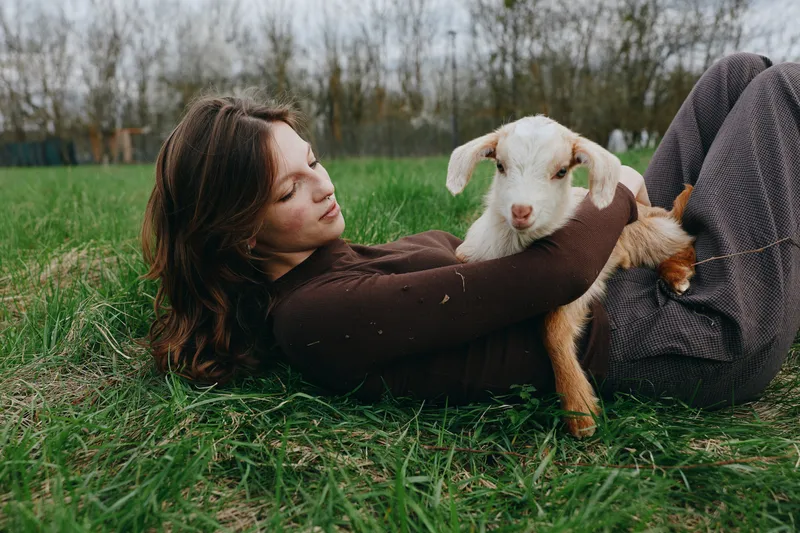 Young woman in brown sweater lies on grass gently holding a white baby goat in her arms in a peaceful outdoor setting during a casual lifestyle moment.