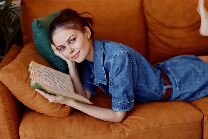 Женщина в джинсовом платье отдыхает на диване, читает книгу, положив ноги наверх, в уютной домашней обстановке