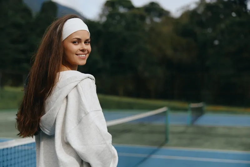 Young cheerful woman with long hair wearing a hoodie and headband smiling on a tennis court in a lush green park setting with a calm background