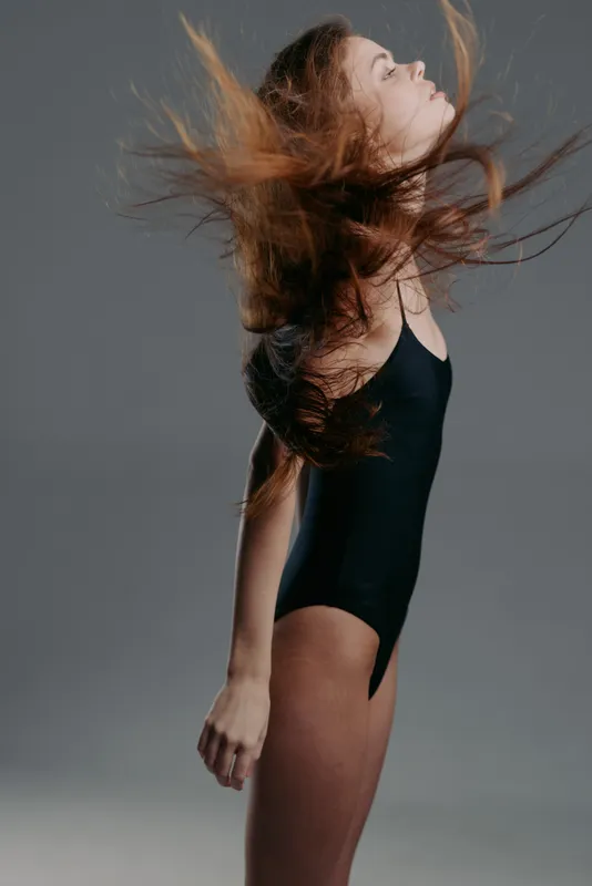 Woman with long hair tossing and flowing in black swimsuit against gray background in motion.