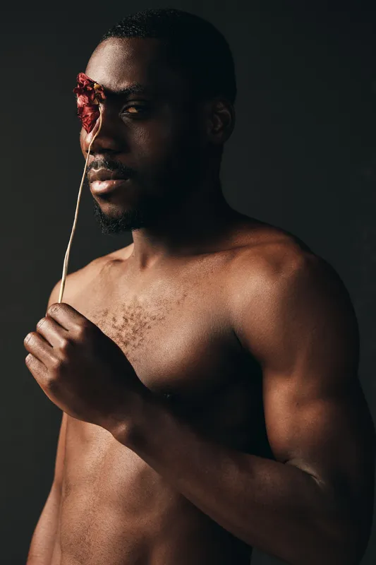 African man with dark skin holding dried flower in front of face, creative conceptual portrait with artistic visual style and bare torso on dark background.