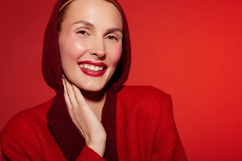 Smiling woman in red hooded sweater poses with hand on cheek against red background. Close-up portrait with natural makeup and joyful expression. Concept of happiness, fashion, and beauty.