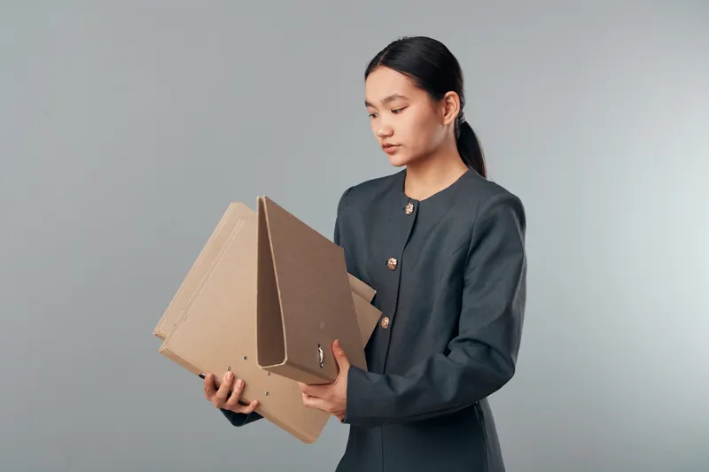 Woman holding cardboard boxes for shipping and delivery with focused expression in studio. Female in dark outfit handling parcels for logistics and packaging process.