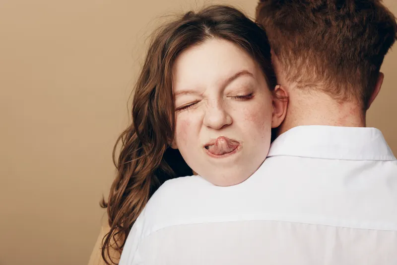 Young woman with closed eyes and playful expression sticks out tongue while hugging man in white shirt. Intimate moment, casual love, and affectionate relationship concept.