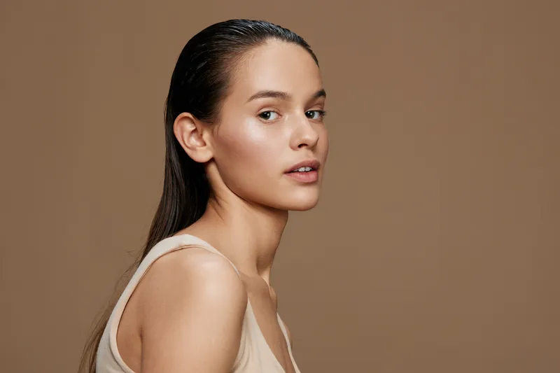A young woman with long straight hair gazing confidently at the camera, wearing a beige sleeveless top against a soft brown background, embodying natural beauty and elegance