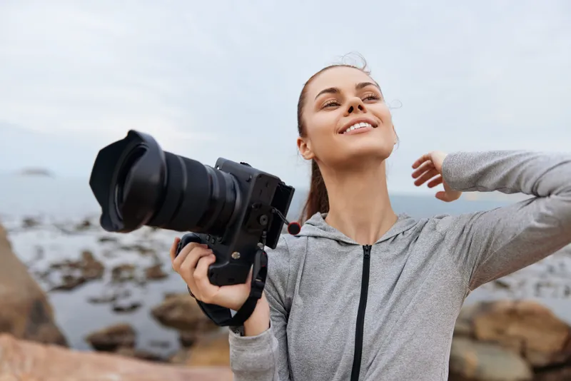 Happy young woman holding a camera outdoors, dressed in casual gray clothing, enjoying the subtle ocean scenery while capturing memories with a smile