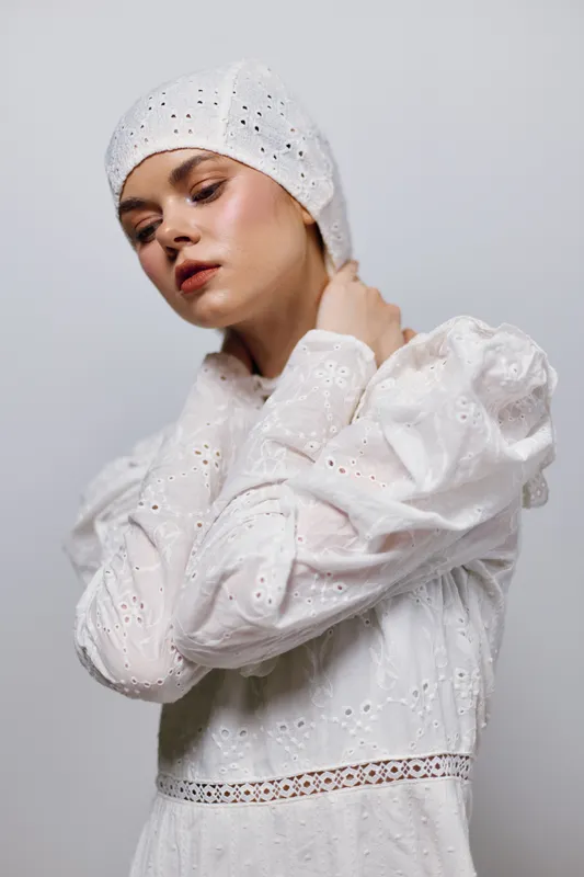 Young model with a serene expression in a white vintage dress and headscarf, showcasing a minimalist style against a soft gray background