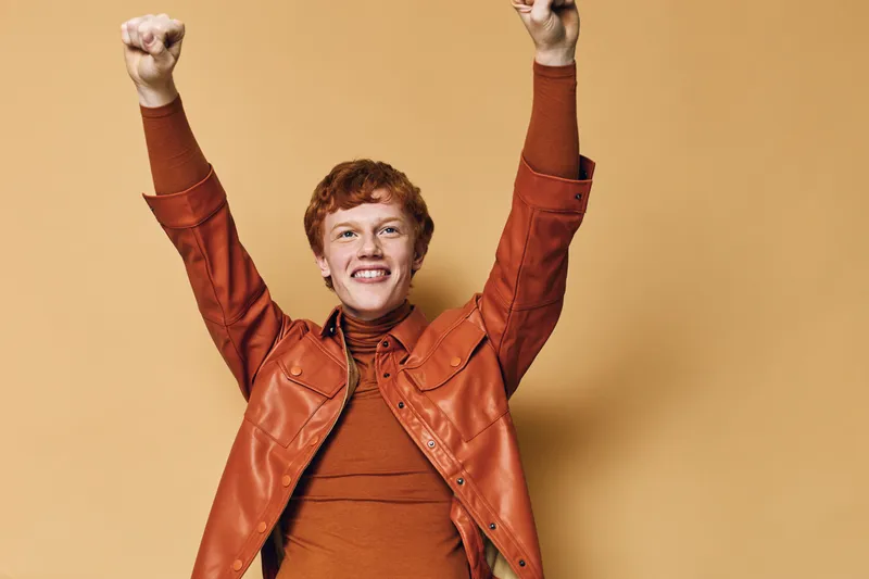 Happy young man with red hair and freckles raising hands up in excitement wearing brown leather jacket and turtleneck on beige background.