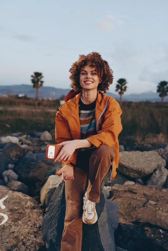 Woman lifestyle outdoors, curly hair, orange jacket, sitting on rocks and holding smartphone during travel in nature, palm trees and mountains in soft daylight, relaxed smiling mood