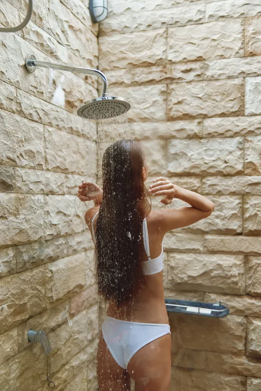 woman showering outdoors in swimsuit with long wet hair against textured stone wall, refreshing summer hygiene routine under rainfall showerhead
