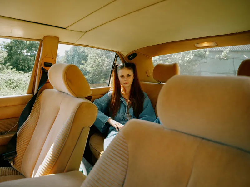 Young woman sitting in a vintage car, showcasing a contemplative expression amidst a warm, minimalist interior with soft beige tones.