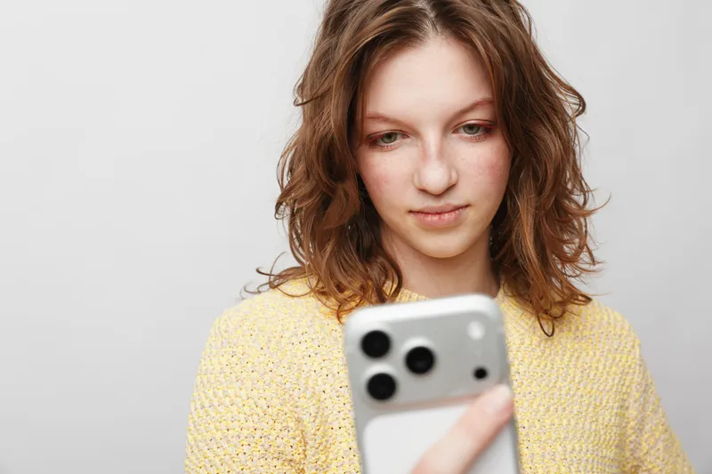 Young woman with curly hair wearing yellow sweater looks at smartphone screen with focused expression. Isolated on light background. Concept of technology, communication, and social media.